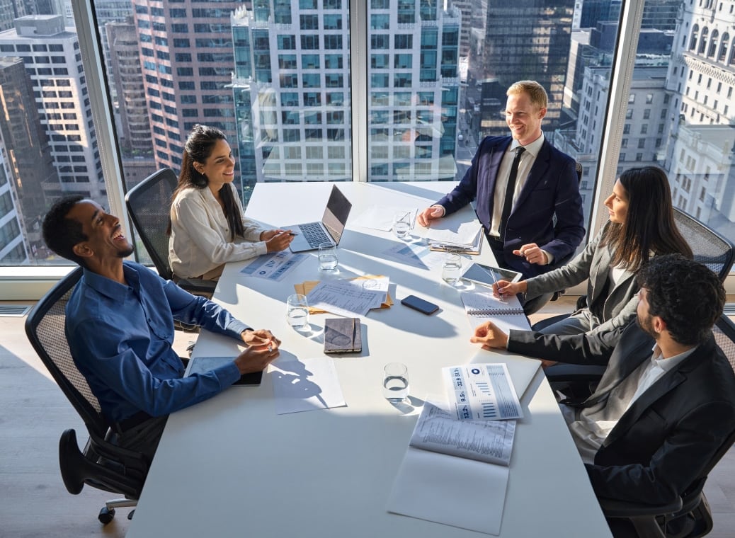 5 business people are sitting at a table in a tall building and talking about business